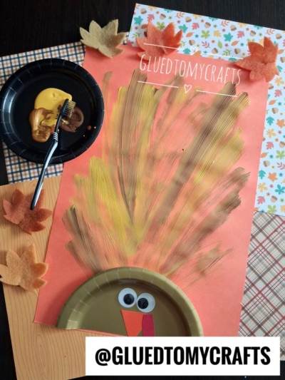 A fun fall crafts for kids project, this turkey craft is made with a painted paper plate, googly eyes, and painted feathers on orange paper. Paint, a fork, and felt leaves are nearby, while plaid and wood surfaces set a cozy background.