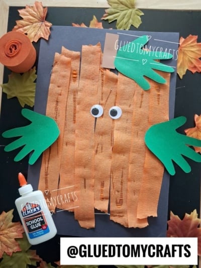 A Halloween craft featuring a Torn Paper Spookley Pumpkin mummy made with orange crepe paper strips, green handprint cutouts for arms, googly eyes, a glue bottle, and fake leaves on a black background.