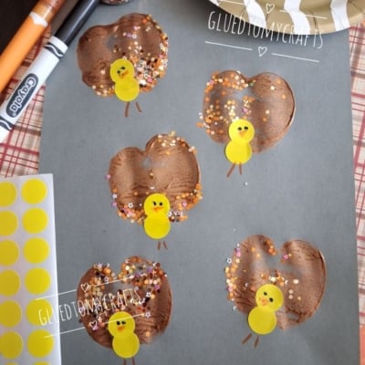 Gray paper with five brown-painted turkey shapes made from handprints, decorated with yellow sticker heads, orange noses, and colorful confetti. Craft supplies and stickers are scattered around.