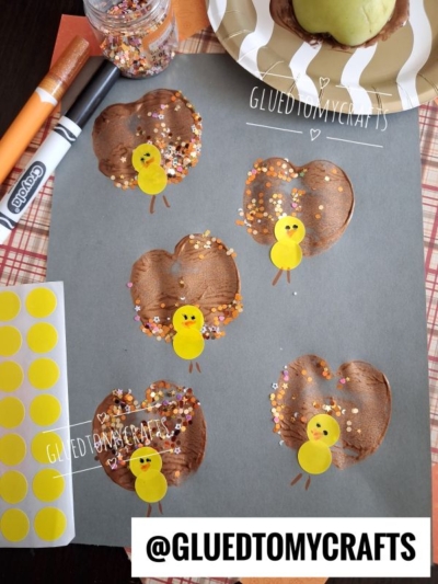 Gray paper with five brown-painted turkey shapes made from handprints, decorated with yellow sticker heads, orange noses, and colorful confetti. Craft supplies and stickers are scattered around.
