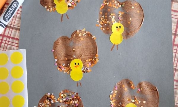 Gray paper with five brown-painted turkey shapes made from handprints, decorated with yellow sticker heads, orange noses, and colorful confetti. Craft supplies and stickers are scattered around.
