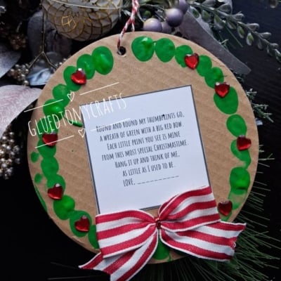 A circular ornament with a wreath of green thumbprints and red heart gems around a poem, finished with a red and white striped bow.