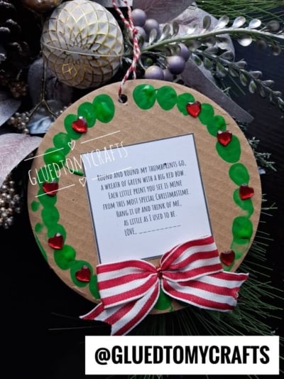 A circular ornament with a wreath of green thumbprints and red heart gems around a poem, finished with a red and white striped bow.