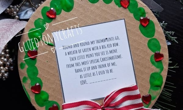 A circular ornament with a wreath of green thumbprints and red heart gems around a poem, finished with a red and white striped bow.