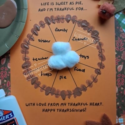 A craft project on orange paper shaped like a pie divided into sections labeled family, friends, toys, food, pie, hugs, teacher, and sister, topped with a cotton ball whipped cream dollop and painted brown crust details.