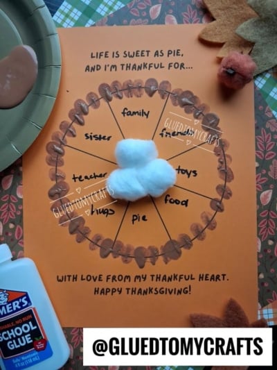 A craft project on orange paper shaped like a pie divided into sections labeled family, friends, toys, food, pie, hugs, teacher, and sister, topped with a cotton ball whipped cream dollop and painted brown crust details.