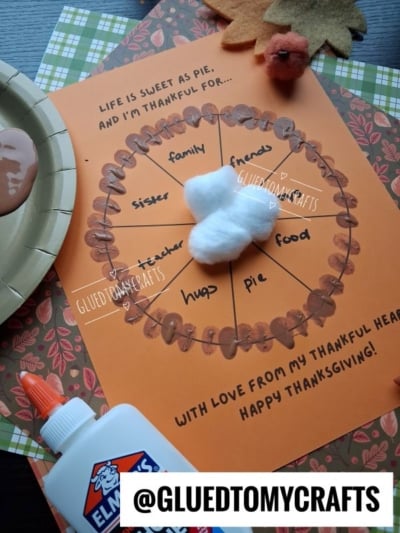A Thanksgiving craft on orange paper shaped like a pie divided into sections, labeled with things to be thankful for, topped with a cotton ball dollop of whipped cream and surrounded by crafting supplies.