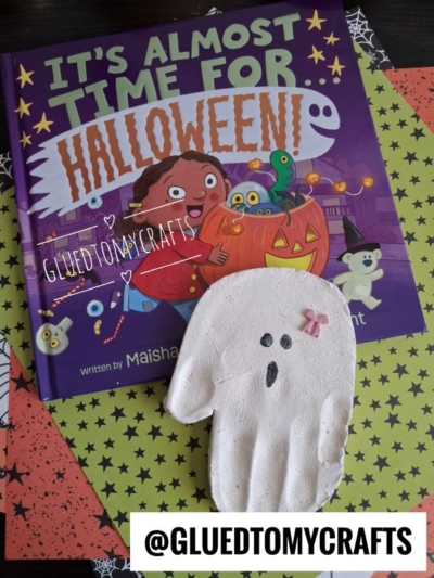 A white clay handprint ghost with a small pink bow resting on the children’s book It’s Almost Time for Halloween, perfect for Halloween crafts for kids.
