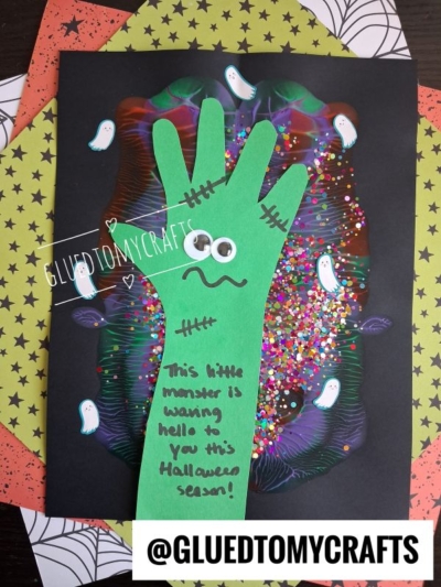 A glittery green handprint cutout with googly eyes and stitches on a black background with ghost illustrations. Text reads, This little monster is waving hello to you this Halloween season! as part of these Halloween crafts for kids.