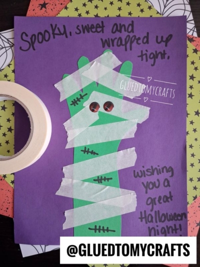 A purple Halloween craft for kids featuring a green construction paper mummy wrapped in masking tape, with button eyes and stitches. It is captioned, Spooky, sweet and wrapped up tight. Wishing you a great Halloween night!.