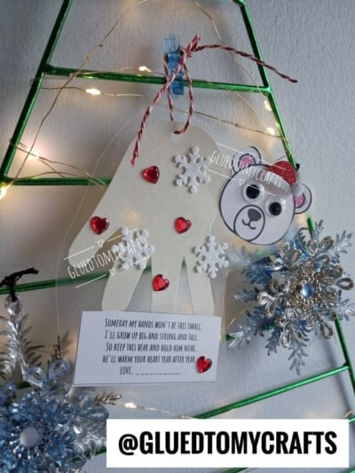 A DIY handprint bear ornament decorated with red heart gems and white snowflakes, hanging on a wire tree with a poem about growing up.