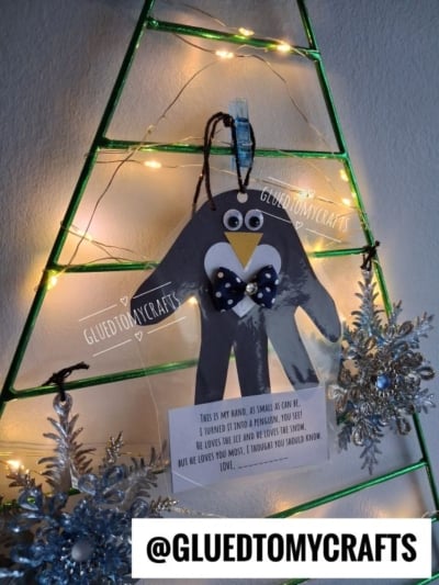 A handprint penguin poem craft wearing a bow tie, hanging on a green wire Christmas tree frame.