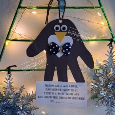 A black paper handprint penguin craft with a poem printed on a card below, hanging on a wire Christmas tree frame with lights.