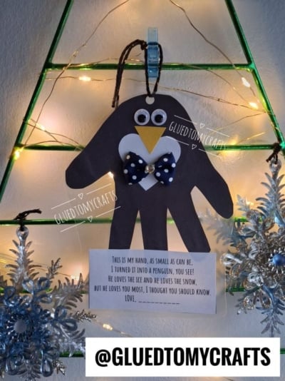 A black paper handprint penguin craft with a poem printed on a card below, hanging on a wire Christmas tree frame with lights.