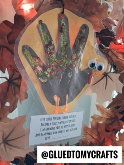 Handprint turkey craft with googly eyes and glitter, featuring a poem about growing fast, hanging in front of a fall leaf wreath.