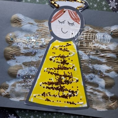 Paper angel craft with glittery yellow wings and a gold halo, set against a dark paper background with snowflake patterns.