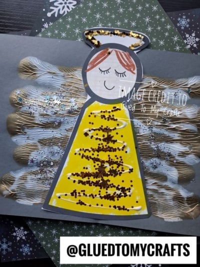 Paper angel craft with glittery yellow wings and a gold halo, set against a dark paper background with snowflake patterns.