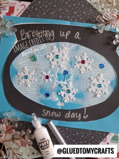 A handmade card featuring a black silhouette of a snowman inside a light blue oval, decorated with white glittery snowflakes, colorful rhinestones, and the text Brewing up a snow day!.