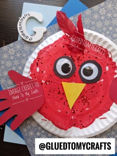 A paper plate craft of a red bird with googly eyes, a yellow beak, and a red paper handprint wing.