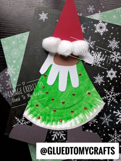 A craft of a paper plate Santa gnome with a red cone hat, white pom-pom trim, and a white handprint beard decorated with red gems.