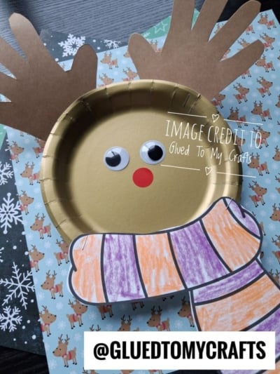A gold paper plate reindeer craft with paper handprint antlers, googly eyes, a red nose, and a coloring-page scarf.