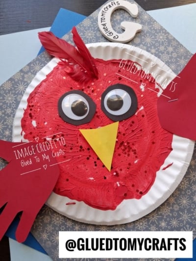 A red paper plate craft shaped like a cardinal with large googly eyes, a yellow beak, and paper handprint wings.