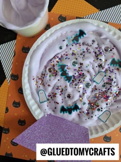 A white paper plate filled with light purple slime decorated with glitter, googly eyes, and paper cutouts of ghosts and bats on a black cat-patterned surface.