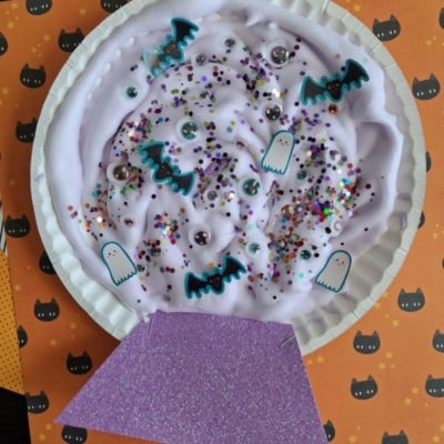 A paper plate craft shaped like a crystal ball, filled with purple slime, colorful glitter, googly eyes, and paper cutouts of ghosts and bats.