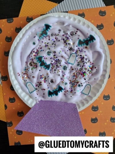 A paper plate craft shaped like a crystal ball, filled with purple slime, colorful glitter, googly eyes, and paper cutouts of ghosts and bats.