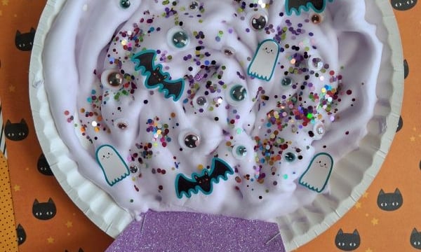 A paper plate craft shaped like a crystal ball, filled with purple slime, colorful glitter, googly eyes, and paper cutouts of ghosts and bats.