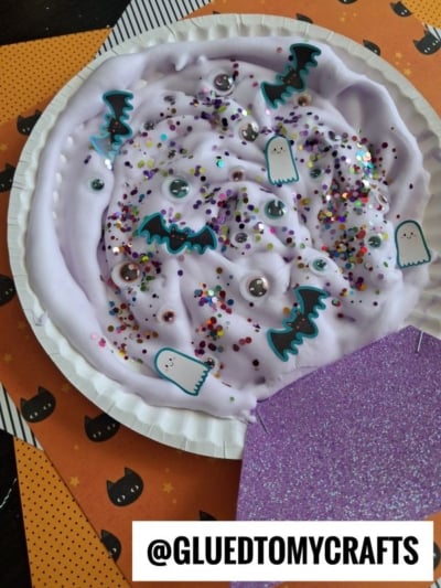 A paper plate filled with light purple slime decorated with googly eyes, glitter, and paper cutouts of ghosts and bats.