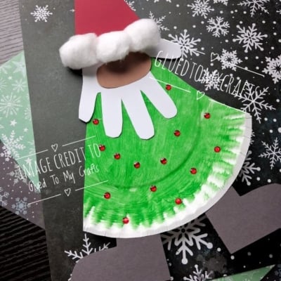 A Christmas craft made from a paper plate, featuring a red hat with cotton balls, a white handprint beard, and a green plate body decorated with red rhinestones.