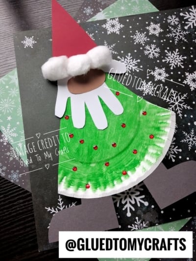 A Christmas craft made from a paper plate, featuring a red hat with cotton balls, a white handprint beard, and a green plate body decorated with red rhinestones.