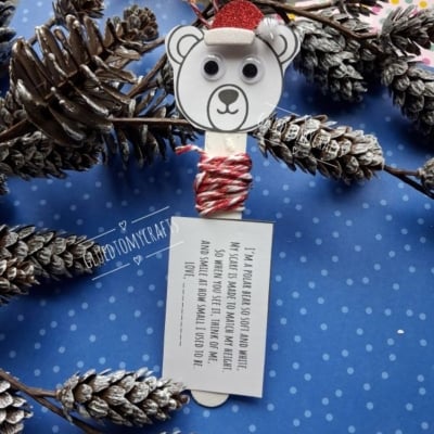 A popsicle stick polar bear craft with a glittery red hat, googly eyes, and a tag with a poem about a polar bear, surrounded by pine cones on a blue polka-dot background.