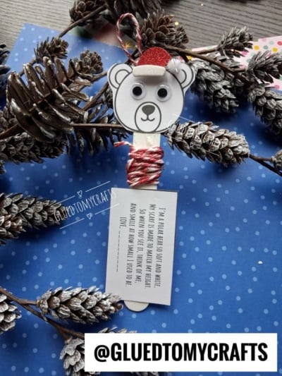 A popsicle stick polar bear craft with a glittery red hat, googly eyes, and a tag with a poem about a polar bear, surrounded by pine cones on a blue polka-dot background.