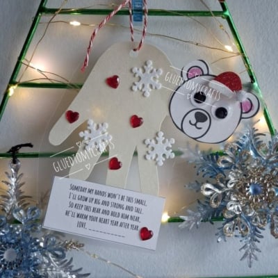 A handmade ornament featuring a paper handprint, a polar bear face, and a poem about growing up, decorated with red hearts and white snowflakes.