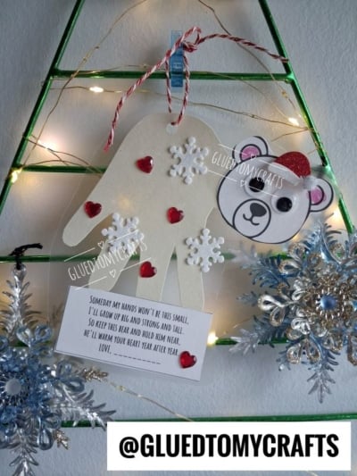 A handmade ornament featuring a paper handprint, a polar bear face, and a poem about growing up, decorated with red hearts and white snowflakes.