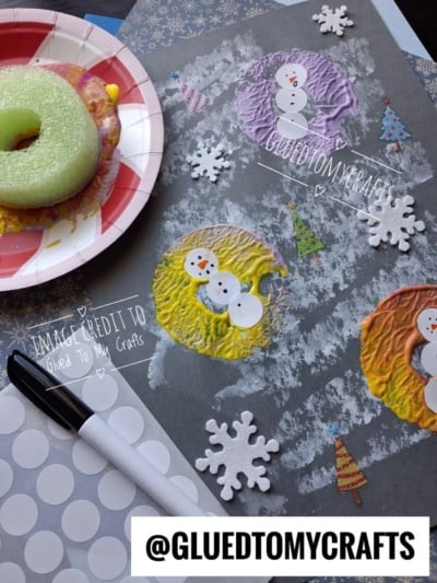 Paper plate snowman craft on gray paper, with a green donut used as a stamp for the body, alongside small snowflake and tree stickers.