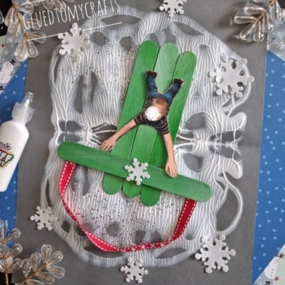 A craft project featuring a paper cutout of a child lying on a sled made of three green craft sticks, accented with white foam snowflakes on a gray background.