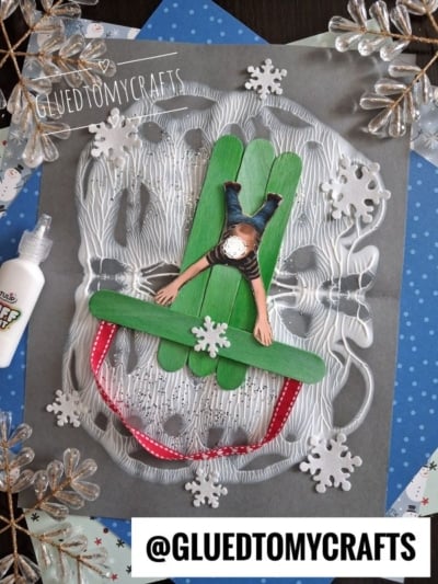 A craft project featuring a paper cutout of a child lying on a sled made of three green craft sticks, accented with white foam snowflakes on a gray background.