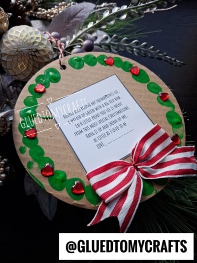 A round craft ornament featuring a painted green wreath with red heart gems and a red-and-white striped ribbon, surrounding a poem about a childs handprint.