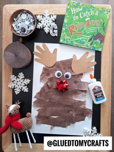A paper reindeer craft on a wooden table surrounded by craft supplies, a childrens book, and a small reindeer figurine.