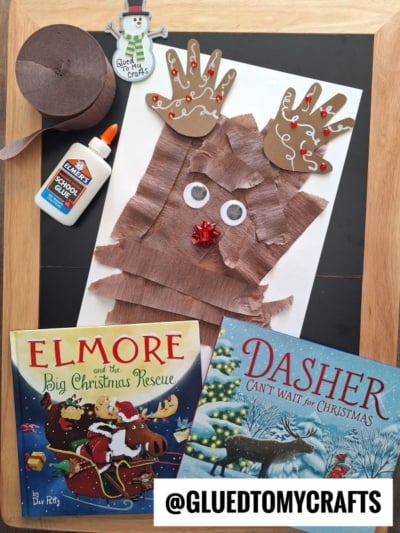 A handmade reindeer craft made from paper handprints and torn brown paper strips, displayed with glue, brown paper, and two Christmas childrens books.