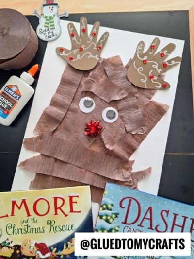 A childs handprint reindeer craft made with brown crepe paper strips, googly eyes, a red bow nose, and cutout handprint antlers decorated with red gems.