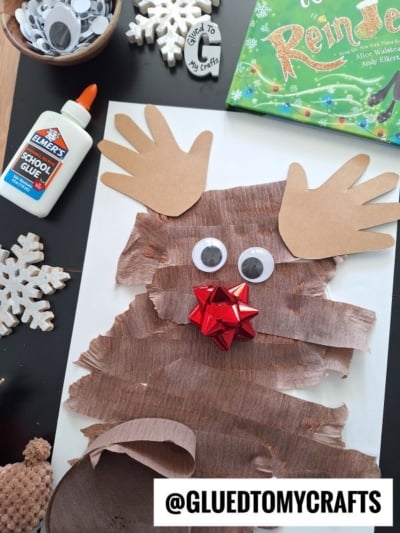 A homemade reindeer craft made of torn brown paper, googly eyes, a red bow nose, and brown paper handprint antlers, surrounded by glue, snowflake cutouts, and a book.