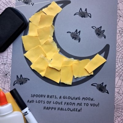 Halloween craft on gray paper featuring a crescent moon made of torn yellow paper, surrounded by small bat thumbprints with hand-drawn wings, and the printed text, Spooky bats, a glowing moon, and lots of love from me to you! Happy Halloween!.