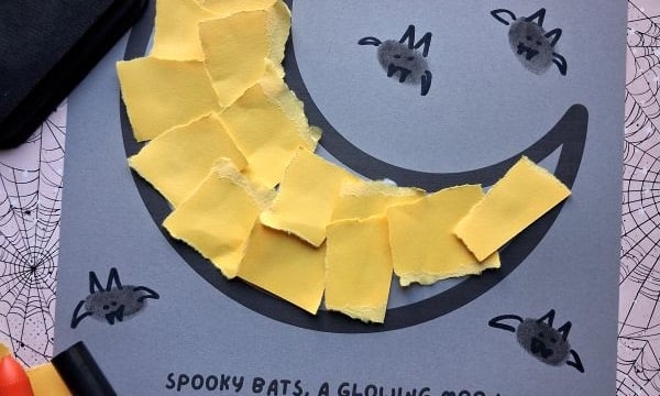 Halloween craft on gray paper featuring a crescent moon made of torn yellow paper, surrounded by small bat thumbprints with hand-drawn wings, and the printed text, Spooky bats, a glowing moon, and lots of love from me to you! Happy Halloween!.