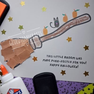 A handmade Halloween card featuring a finger-painted broomstick with a thumbprint cat and pumpkins, decorated with gold star stickers and the text This little broom was made purr-fectly for you! Happy Halloween!.