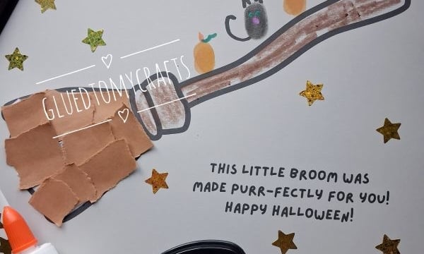 A handmade Halloween card featuring a finger-painted broomstick with a thumbprint cat and pumpkins, decorated with gold star stickers and the text This little broom was made purr-fectly for you! Happy Halloween!.
