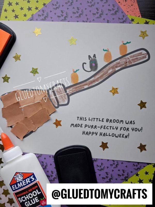 A handmade Halloween card featuring a finger-painted broomstick with a thumbprint cat and pumpkins, decorated with gold star stickers and the text This little broom was made purr-fectly for you! Happy Halloween!.